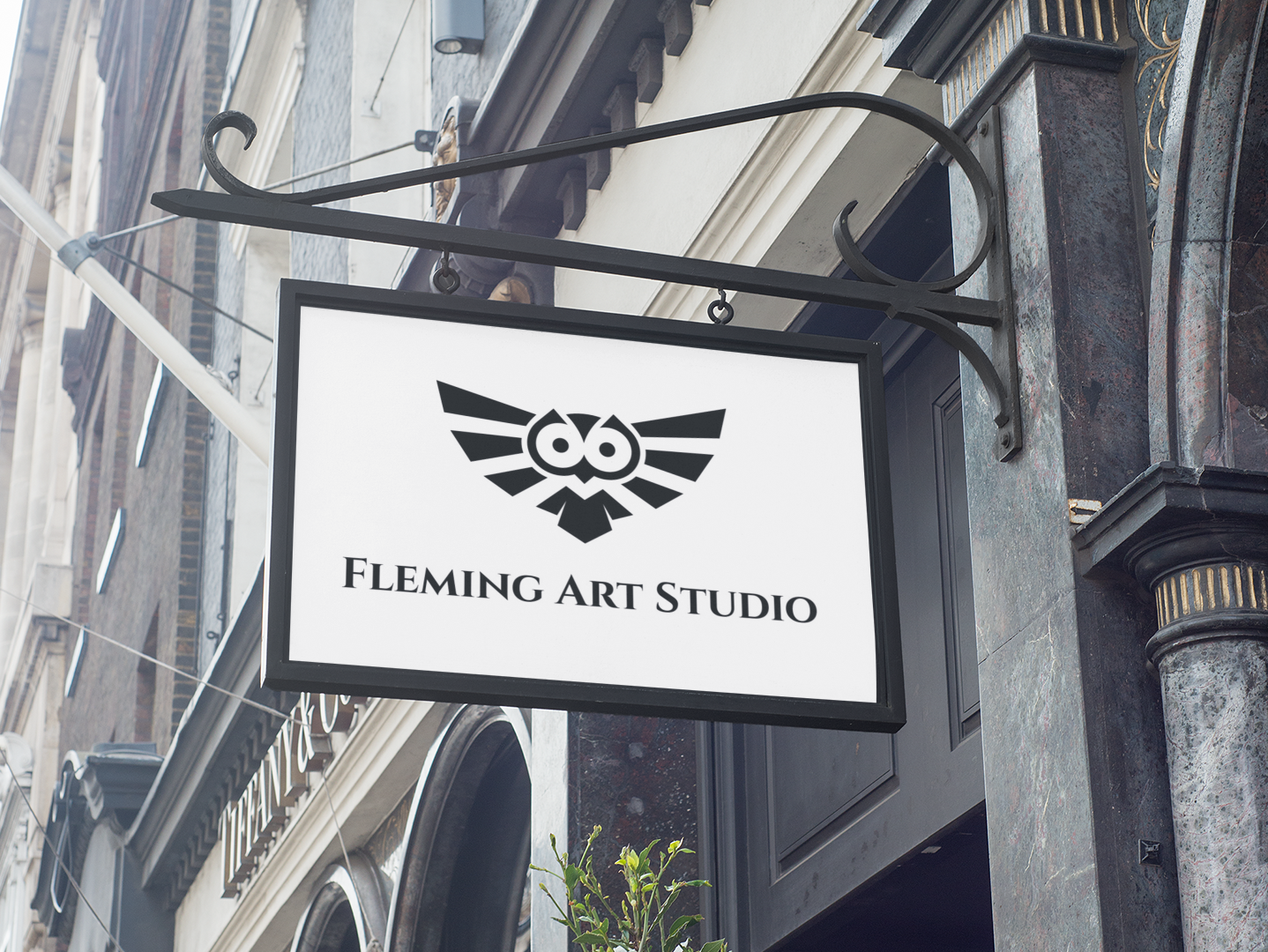 A building with a white rectangular sign attached that features the logo for Fleming Art Studio consisting of a black owl with spread wings and the words Fleming Art Studio underneath it